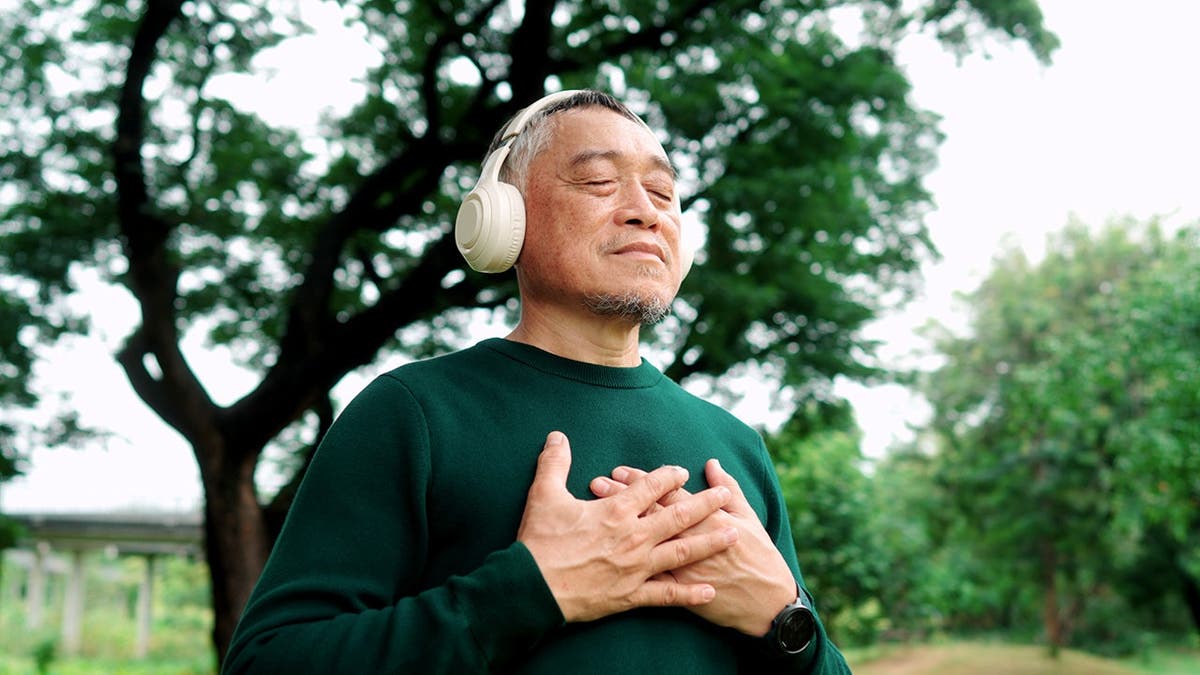 A man wearing headphones smiles and breaths deeply with his hands on his chest outside among trees.