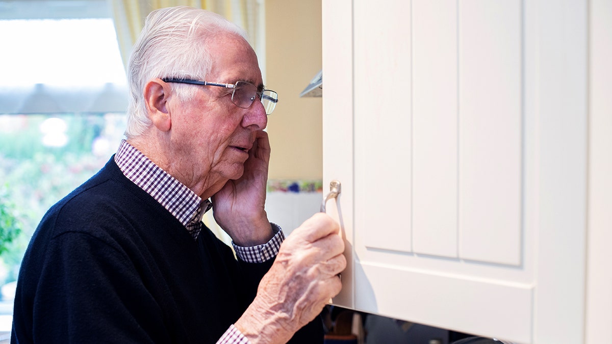 An older man looking confused while glancing inside a cupboard at home