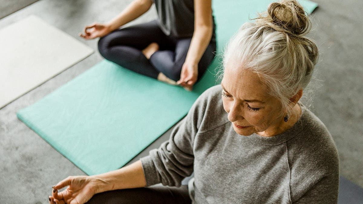 An older woman with gray hair sits on a yoga mat in a yoga pose.