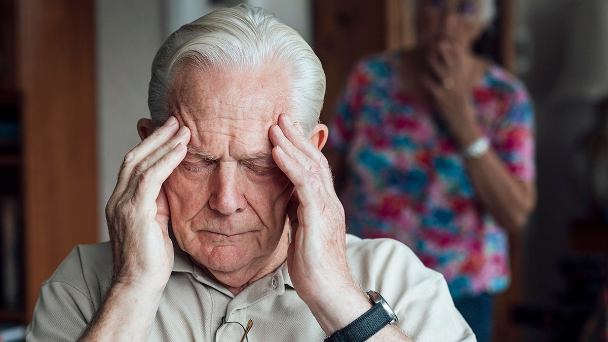 Senior man with Alzheimer's holding his forehead in a living room with wife looking worried behind him