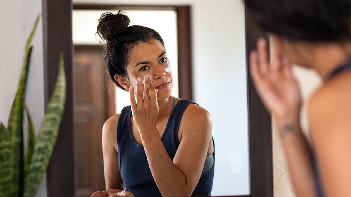 Woman applying white sunblock to her face while looking at bathroom mirror