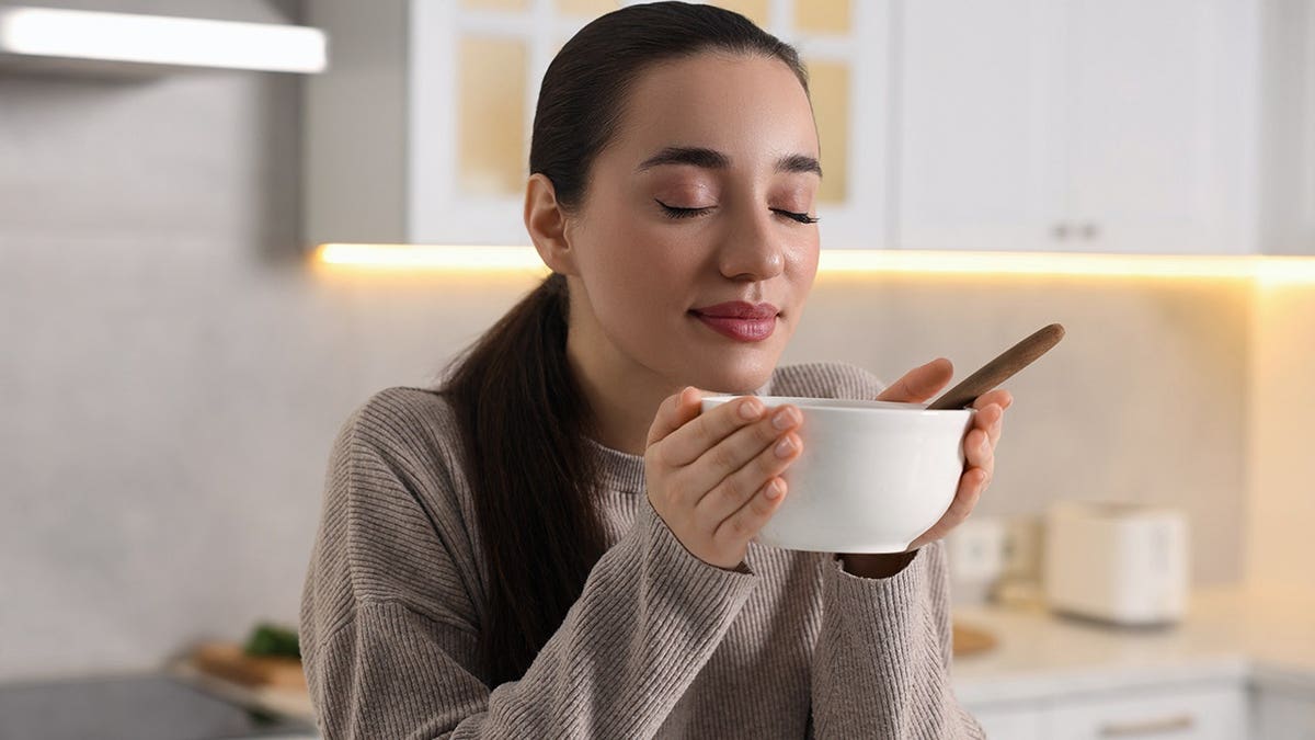 Woman holding a bowl of soup in a kitchen, eyes closed taking in smell.