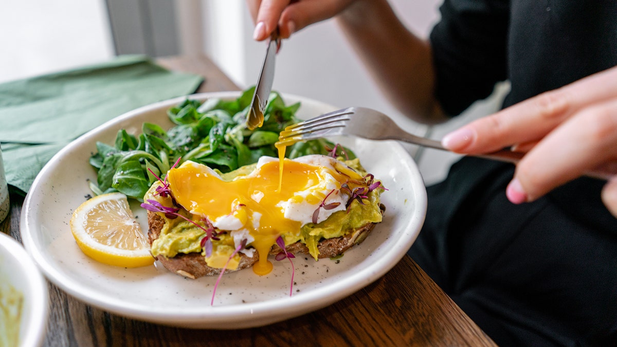 Young woman eating avocado toast topped with poached eggs and salad in a cafe.