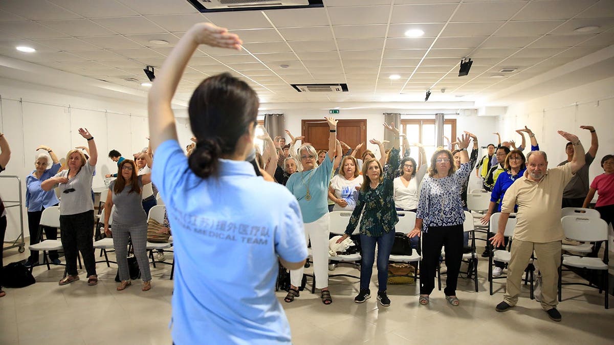 A woman stands in front of a class as she teaches them baduanjin.