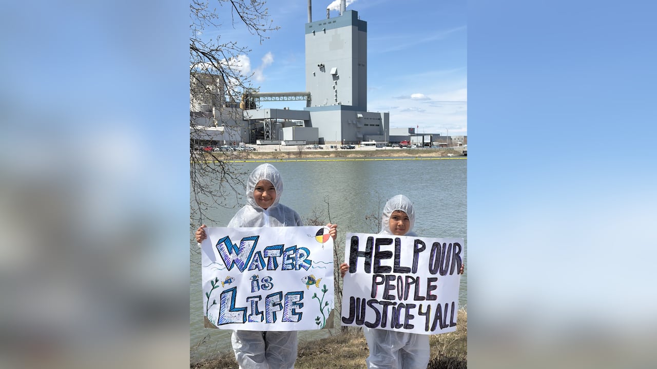 Two young people wearing hazmat suits are seen holding signs outside.