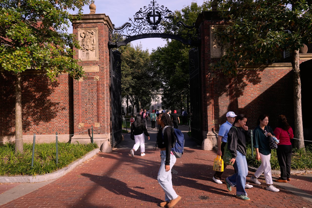 College-aged students walk past a brick wall and iron gate in a leafy campus setting.