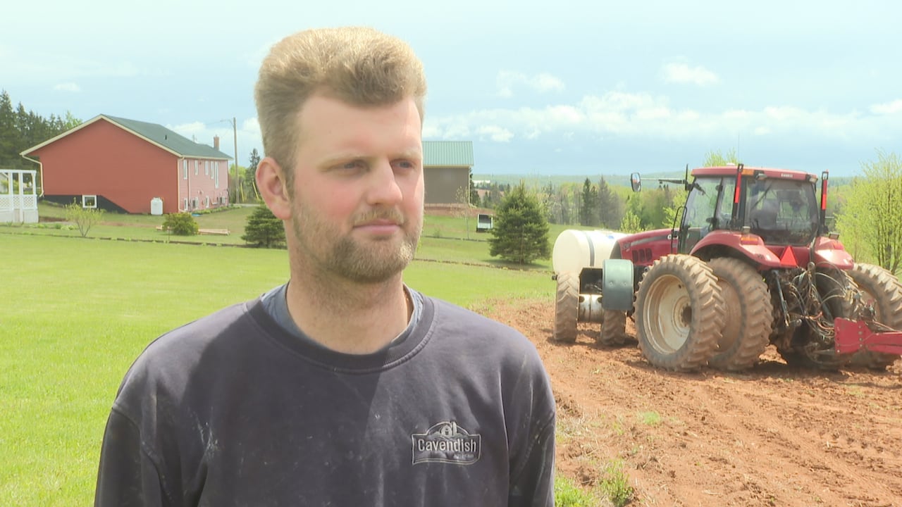 A blonde haired man in a dark sweatshirt stands in front of farm equipment