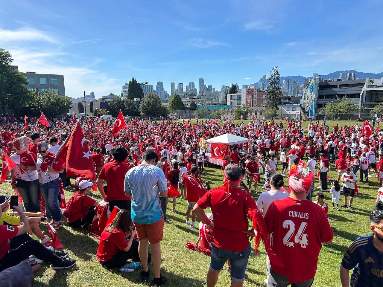 A large crowd of people wearing red and carrying Turkish flags fill a park on a sunny day with the Vancouver skyline visible in the background.