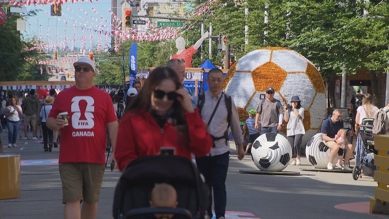 People walk through a pedestrian zone decorated with FIFA-themed displays, including a large soccer ball installation. A man in a red FIFA Canada shirt is seen among pedestrians.