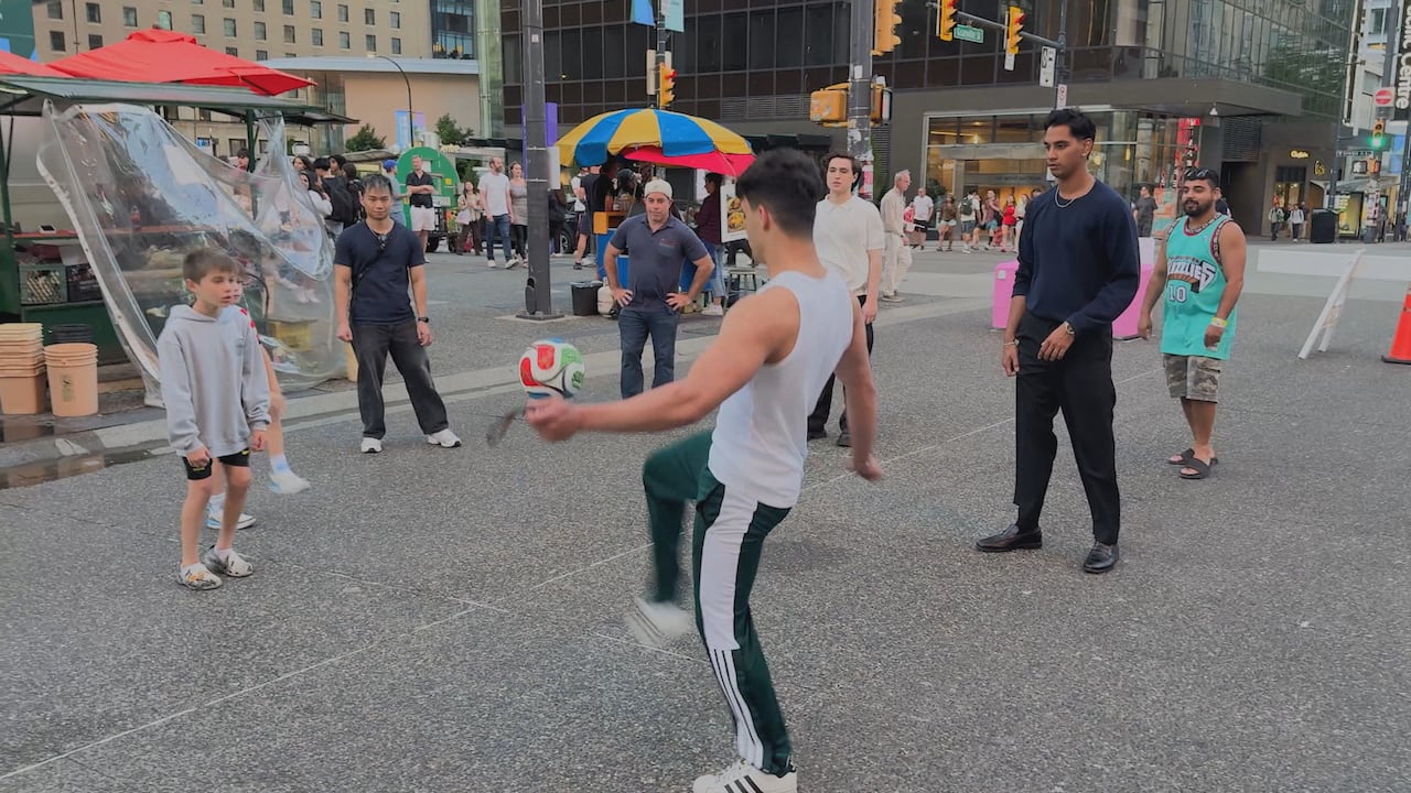 Fans play soccer in a downtown pedestrian zone. Spectators and passersby watch nearby, with food stands and city buildings in the background.