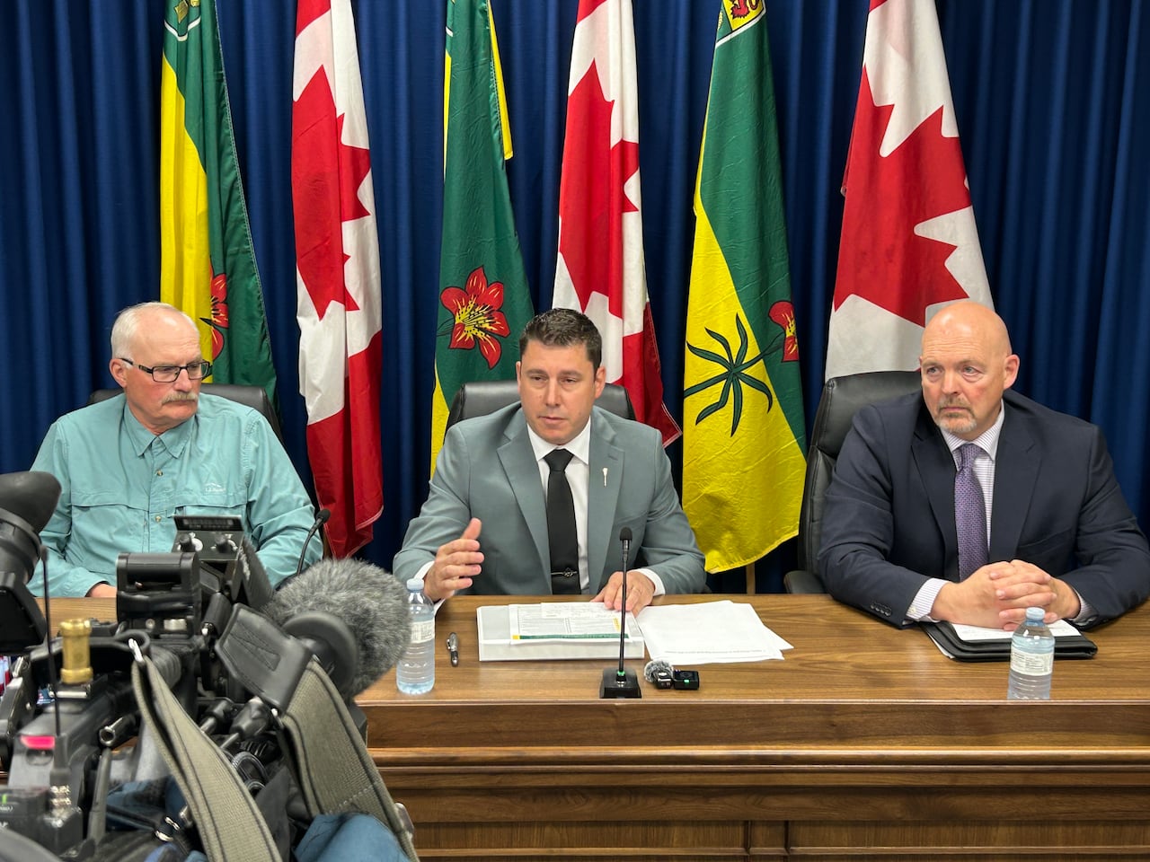 three men sit at a desk at a news conference with Canadian and Saskatchewan flags behind them