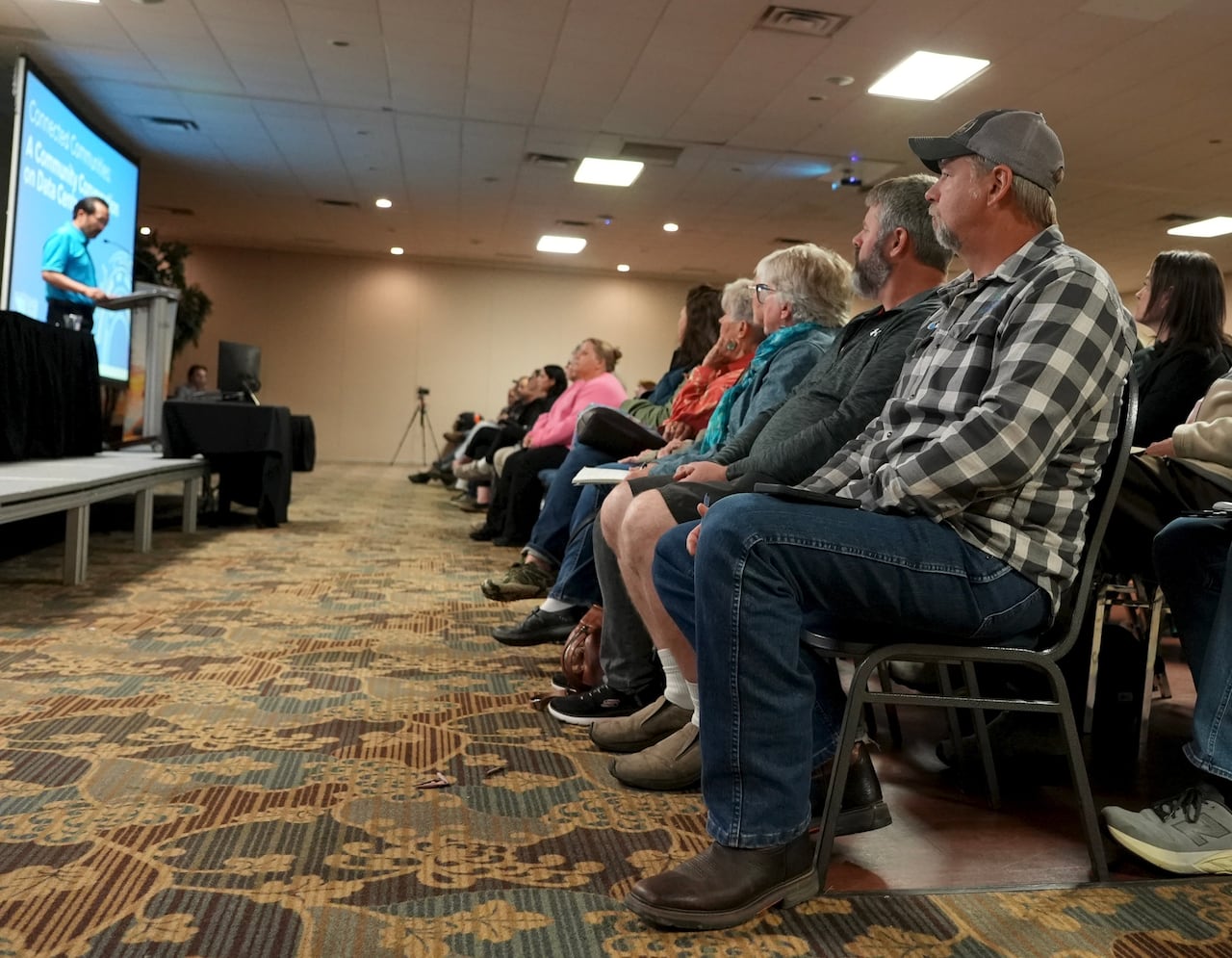 A man in jeans, a checkered shirt and ball cap sits and the end of a long row of seated people who are listening to a man in a blue shirt speak at a podium.