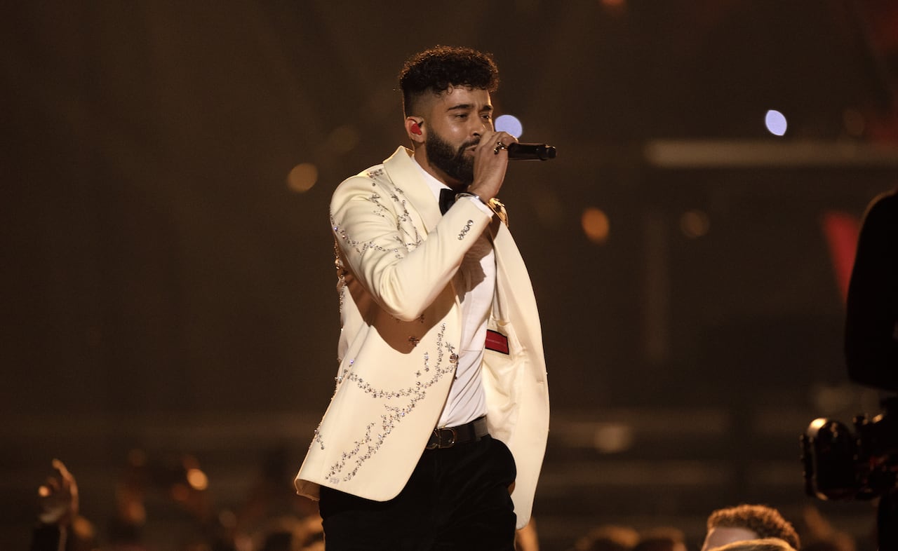 A man with light brown skin and a black beard and black hair wears a white tux-style jacket with gold bling on the shoulders holds a microphone.