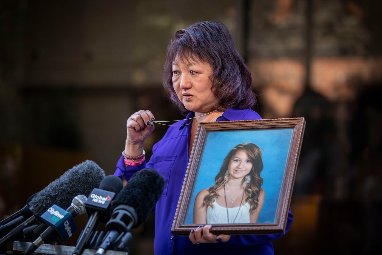 A woman holds out her necklace in one hand while holding a framed picture of a young girl in the other while speaking in front of several microphones.