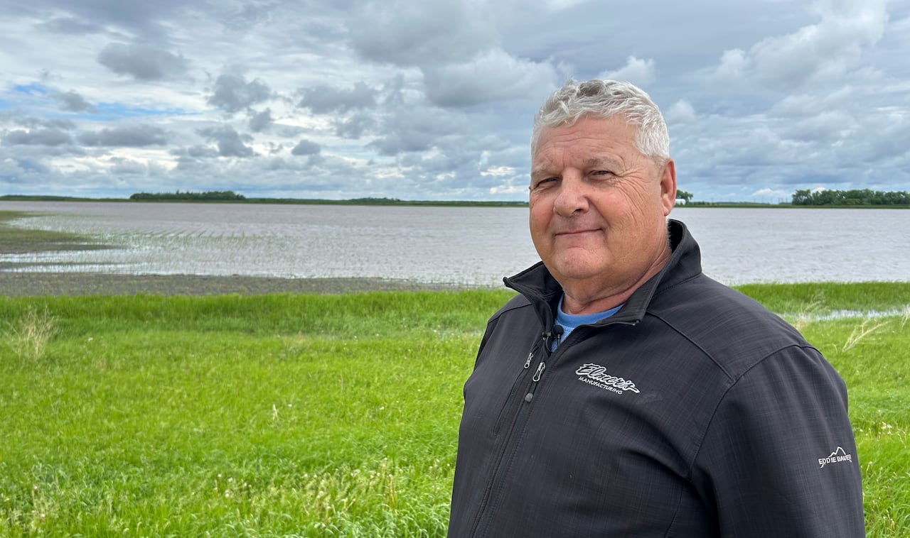 A man with white hair wears a black windbreaker jacket and stands in a green field with water in the background