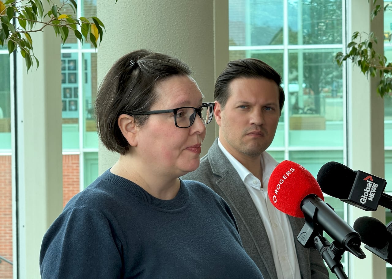 A woman in a blue sweater and glasses speaks in front of news microphones as a man in grey suit with no tie looks on.