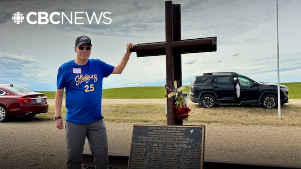 A small crowd stands near an iron cross with a Canadian flag flapping in the background