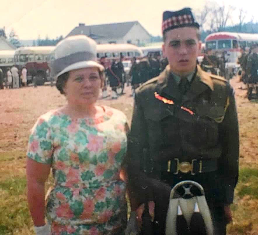 A woman and a teenage boy are standing together. The boy is in military uniform and the woman looks maternal and is wearing an old-fashioned hat.