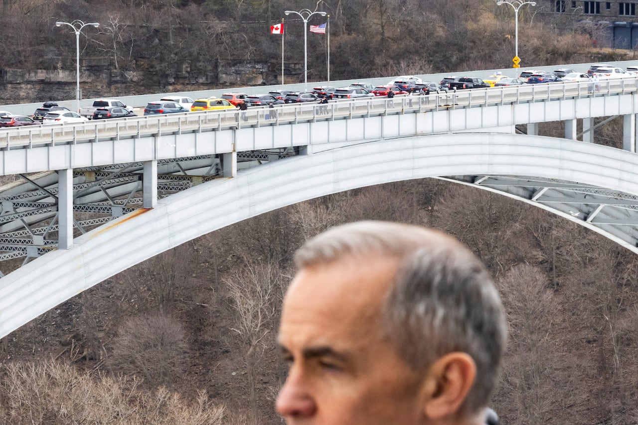 A man with grey hair is seen with a bridge filled with car traffic and a Canadian and American flag in the background.