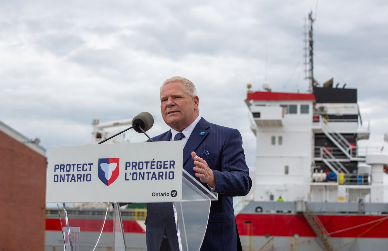A person is seen speaking at a podium. There is a large freighter behind them.