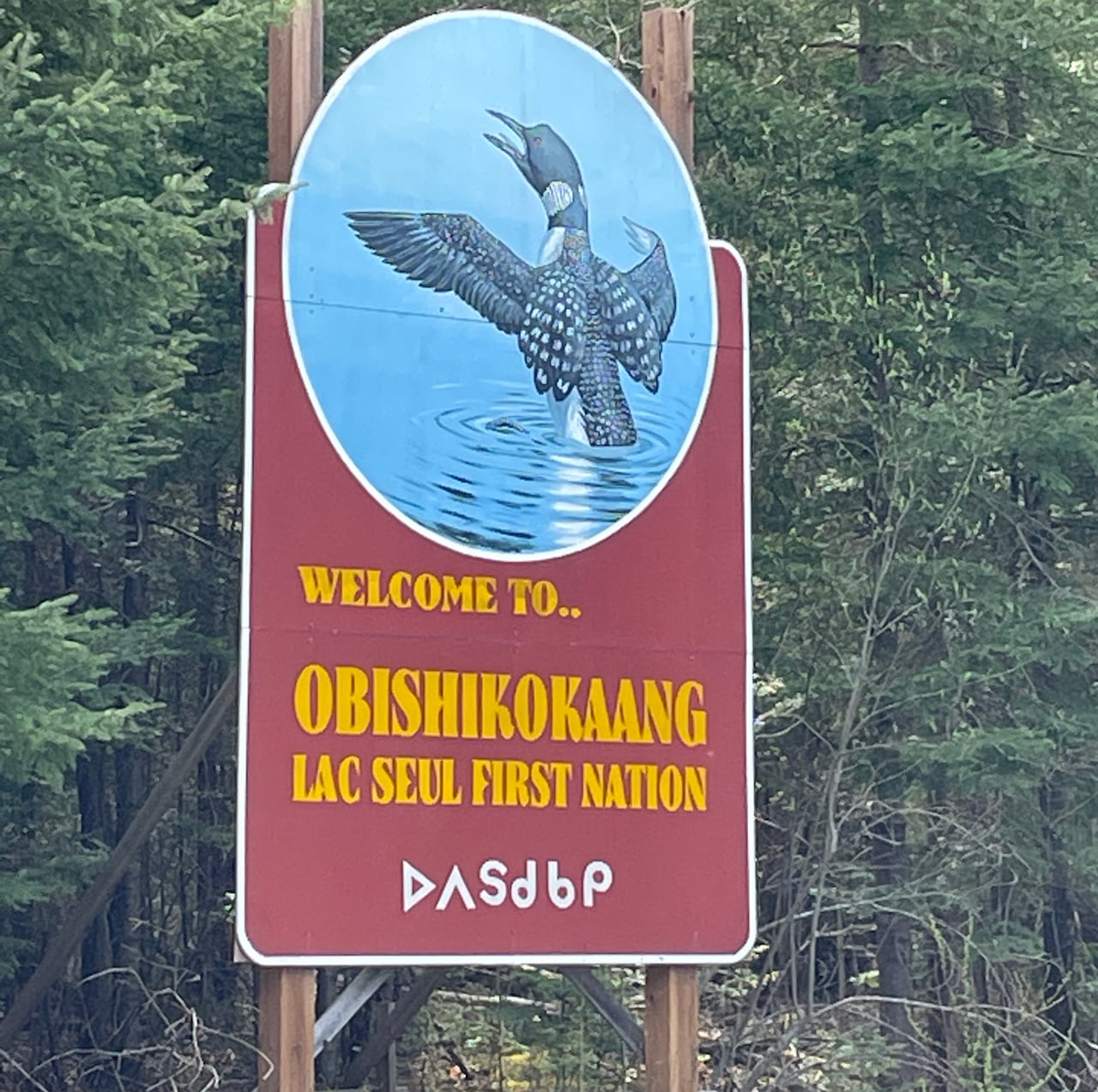 A sign bearing a painted image of a loon rising from water, and the words Welcome To Obishikokaang Lac Seul First Nation.