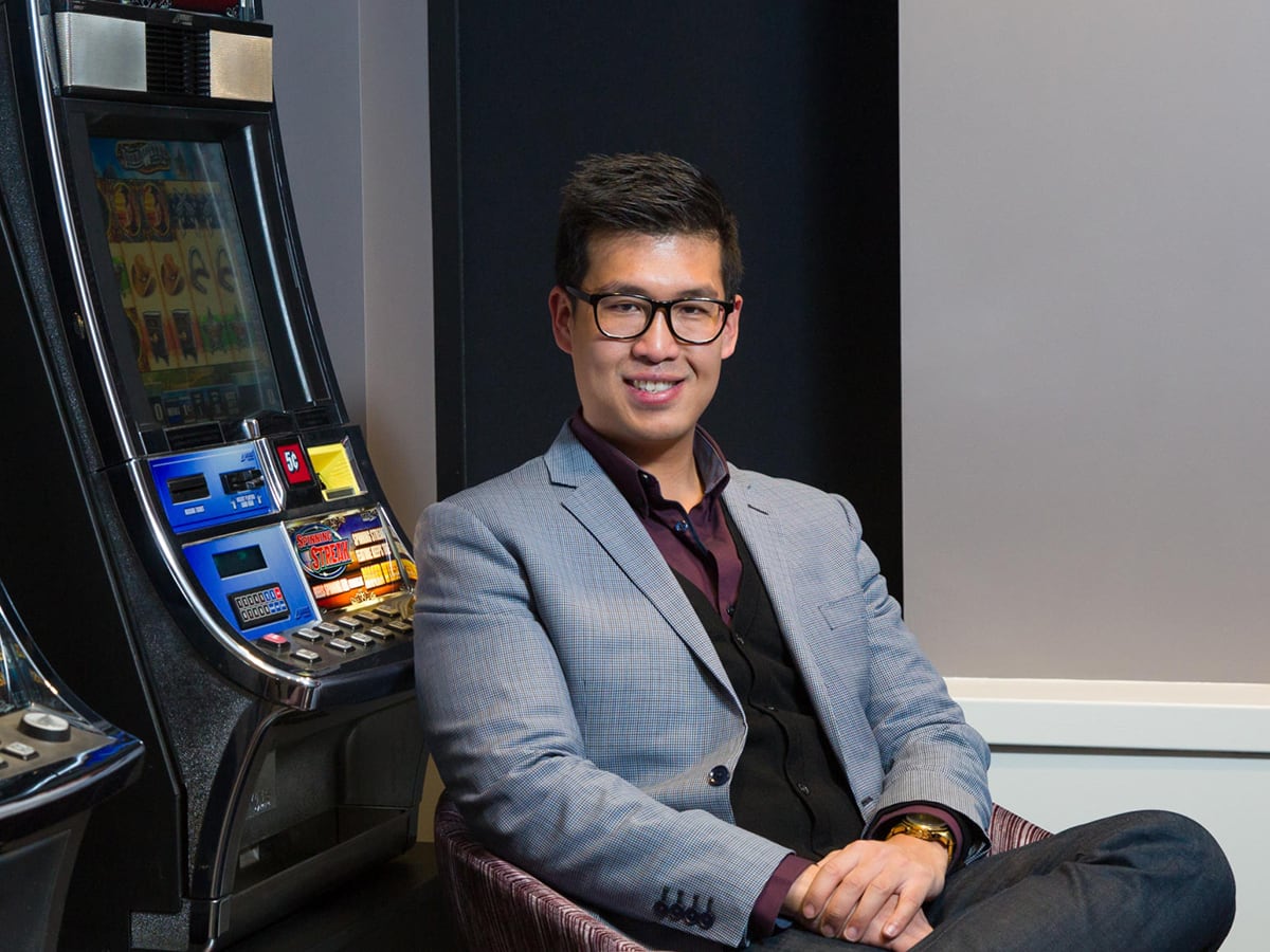 An Asian man wearing glasses, a burgundy shirt and silver check blazer smiles and sits in front of gambilng terminal.