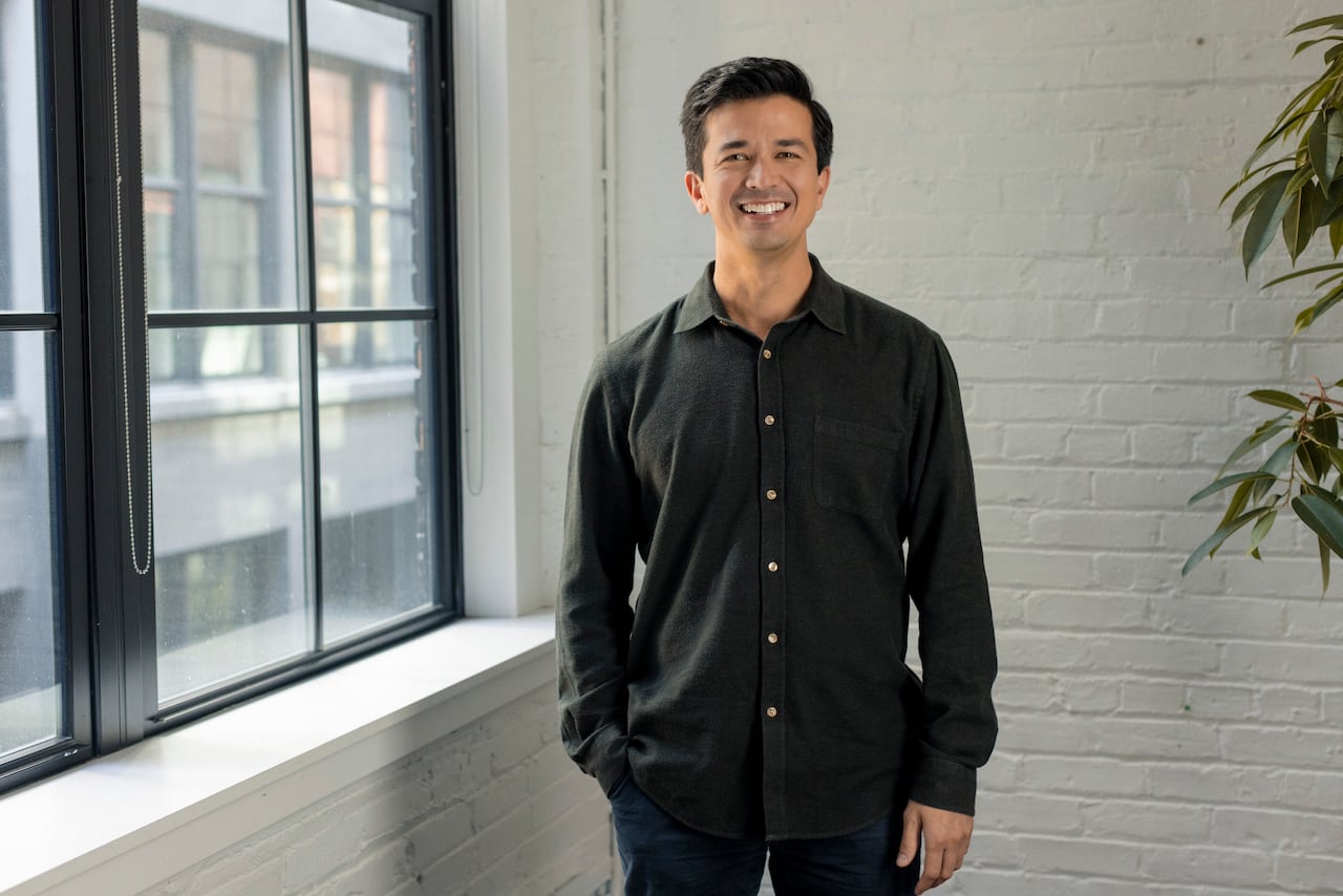 A cean shaven white man with short dark hair wearing a green long sleeve button up shirts stands in front of white brick wall near a large window.