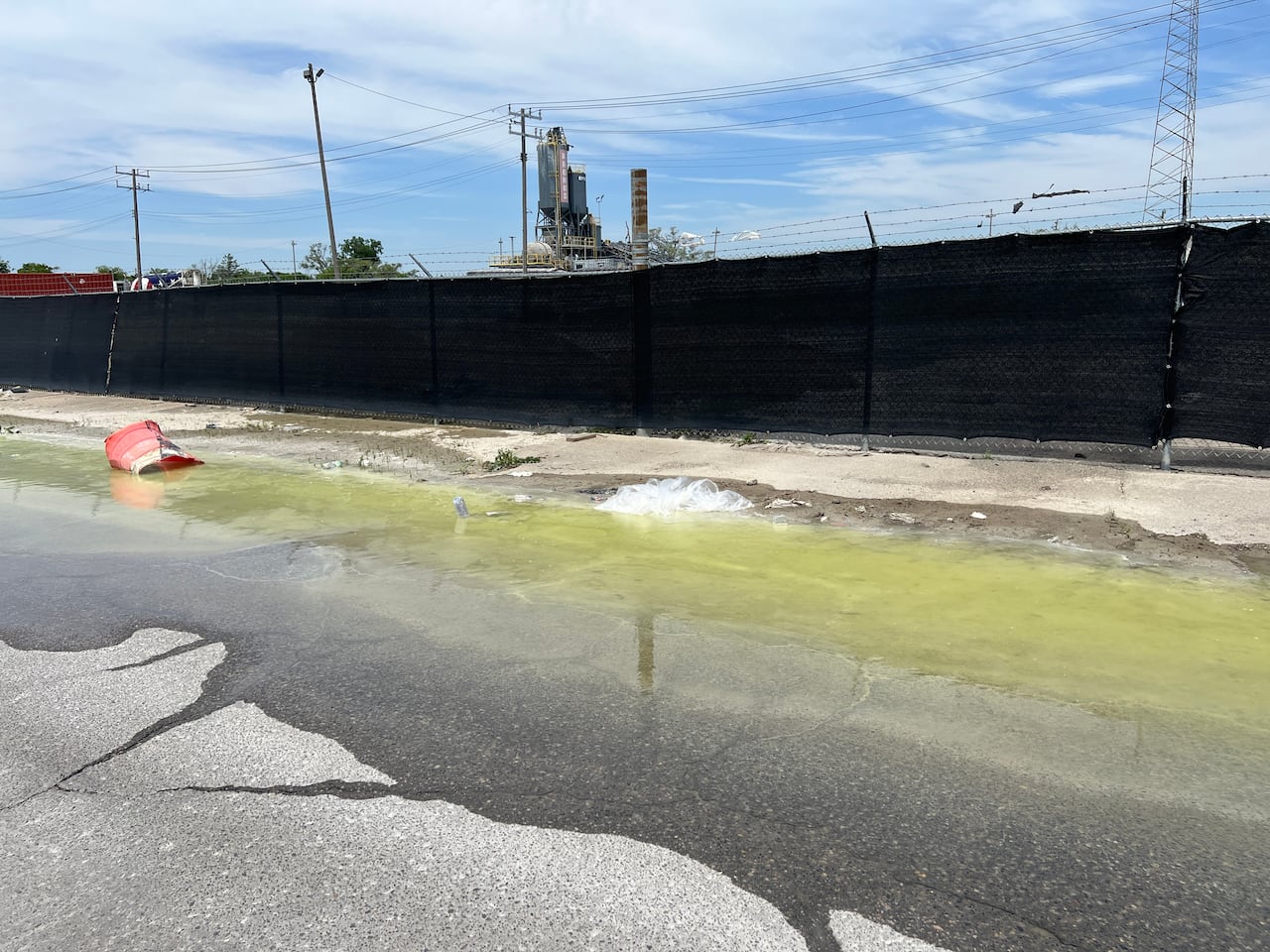 A large green-hued puddle on a street with garbage in front of a concrete facility