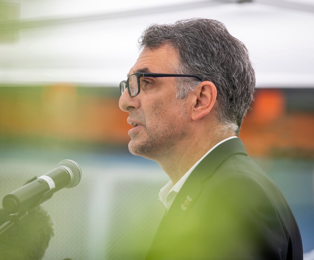A man wearing a suit and glasses is seen in profile speaking to a mic.