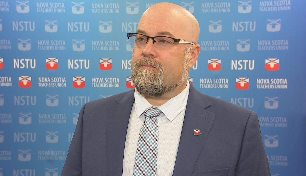 A bald man with glasses and a beard in a suit and tie stands in front of a blue banner with NSTU written across it.