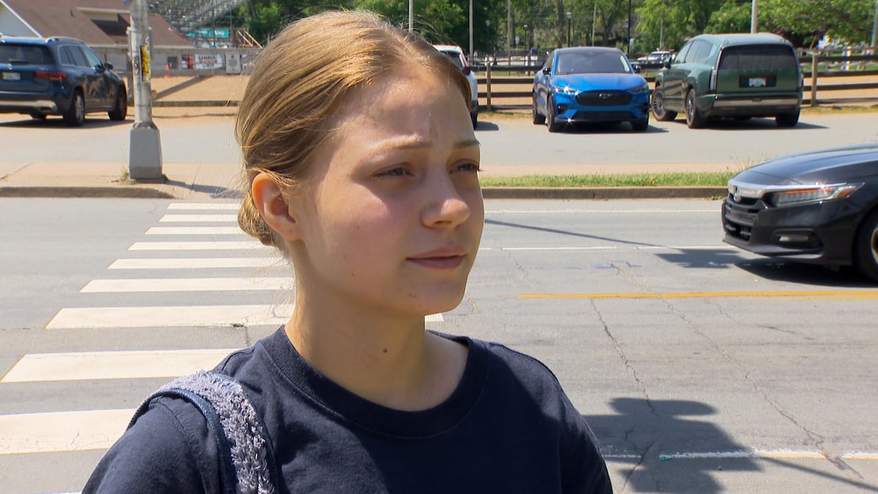 A grade 12 girl wears her hair in a bun. She wears a black T-shirt and is standing in front of a cross walk.