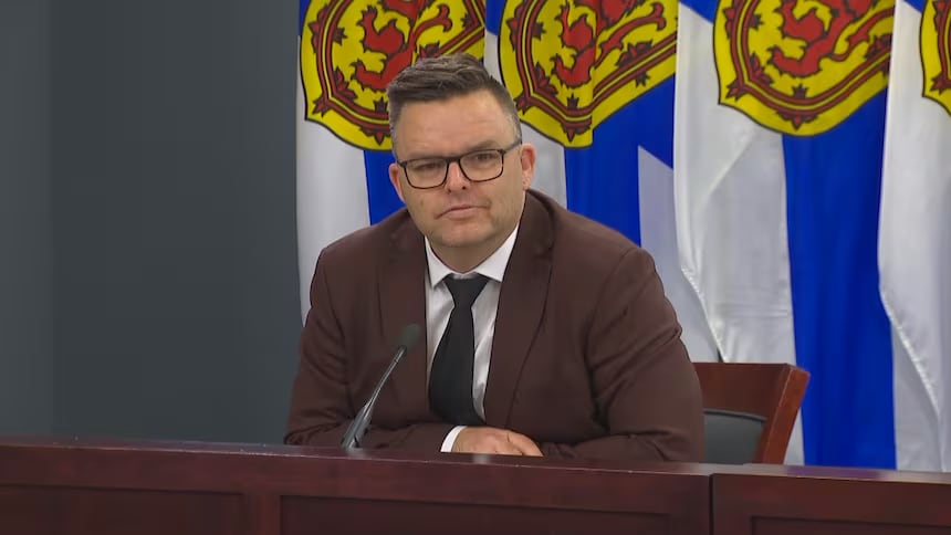 A man wears glasses and a brown suit. He sits at a podium in front of Canadian and Nova Scotia flags.