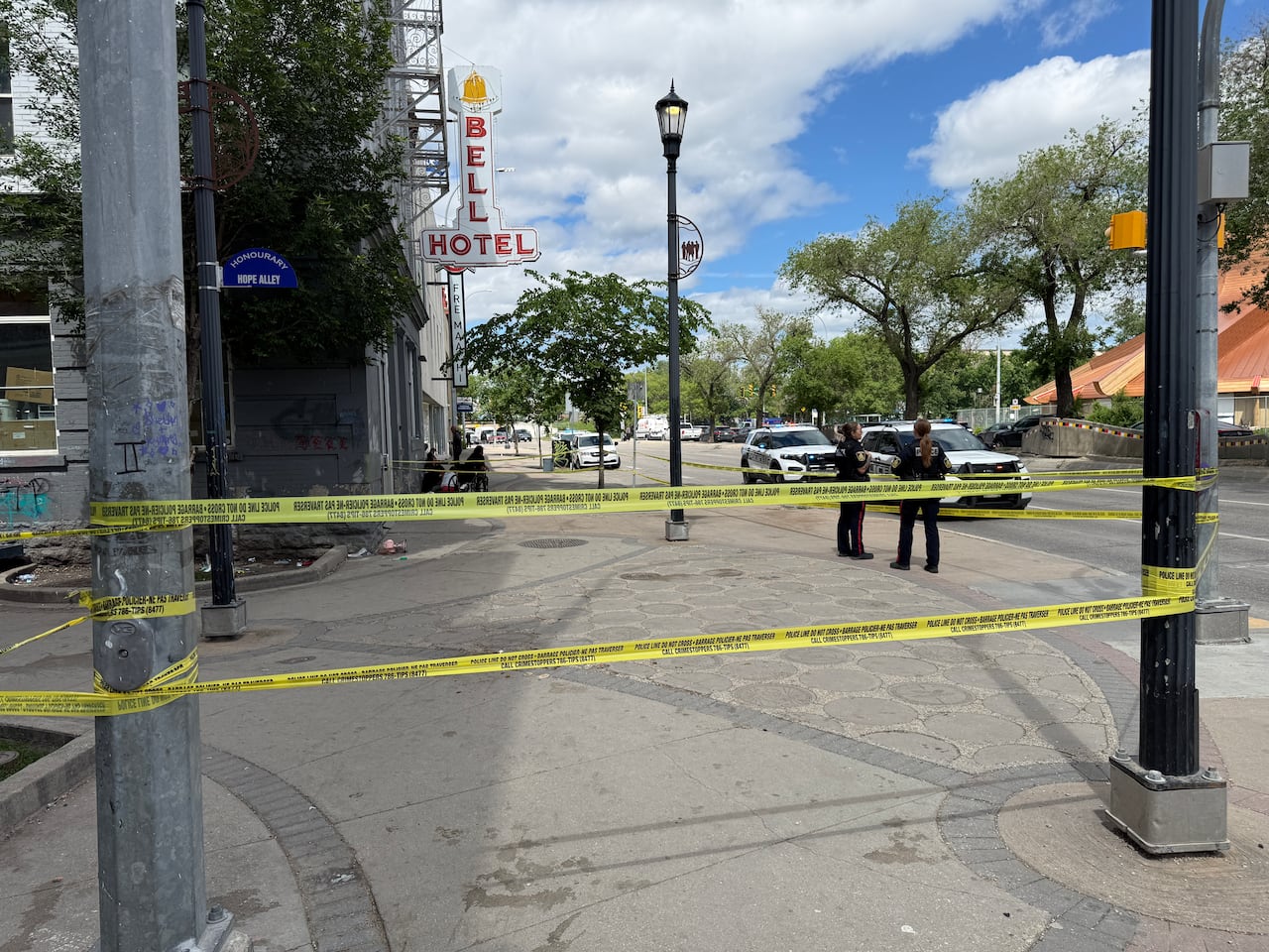 Police tape across on a pone area by a street. There is a sign that says 'Hope Alley.' One building behind the tape has a sign that says 'Bell Hotel.' Police officers and cruisers can be seen in the background.