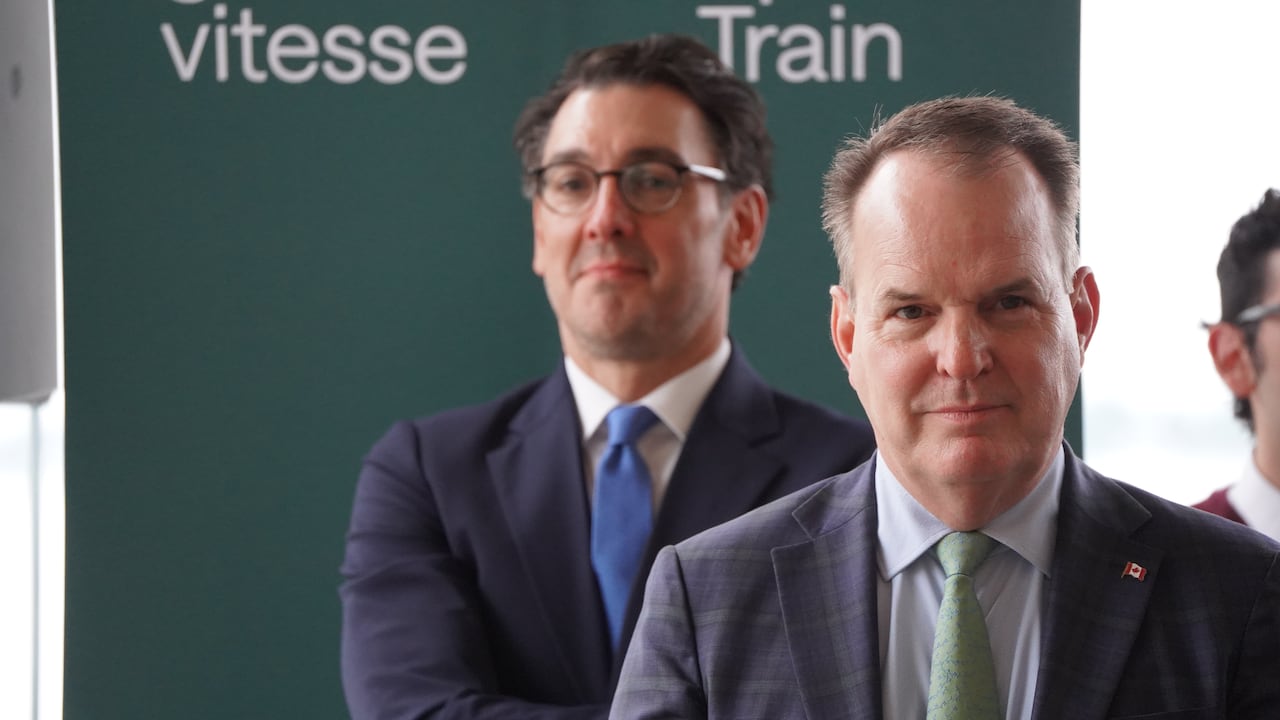 A man with thinning hair and a green tie stares forward. Behind him is a man with glasses and a blue tie standing next to a banner that says "Train."