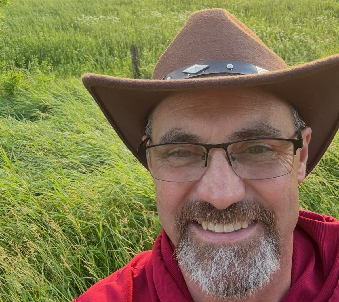 A man in a cowboy hat, with glasses and a goatee, smiles while standing in a grassy field at sunset.