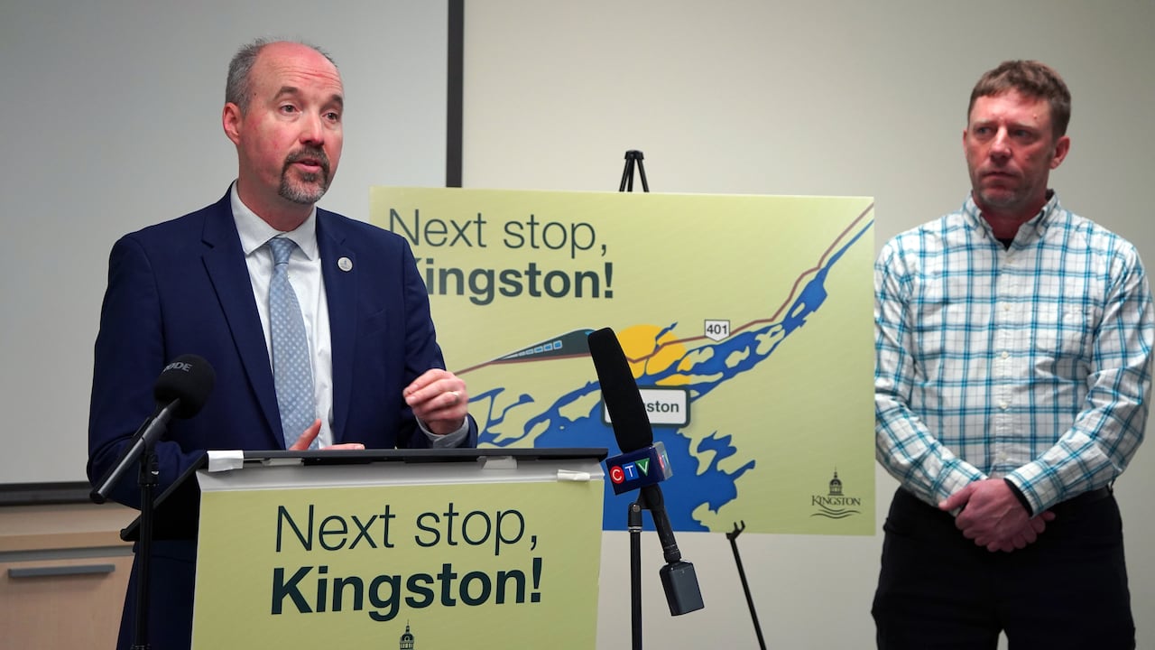 A man with short brown hair and a goatee stands behind a podium with the words "Next stop, Kingston!" on it. A sign behind him bares the same message and a picture of a train.