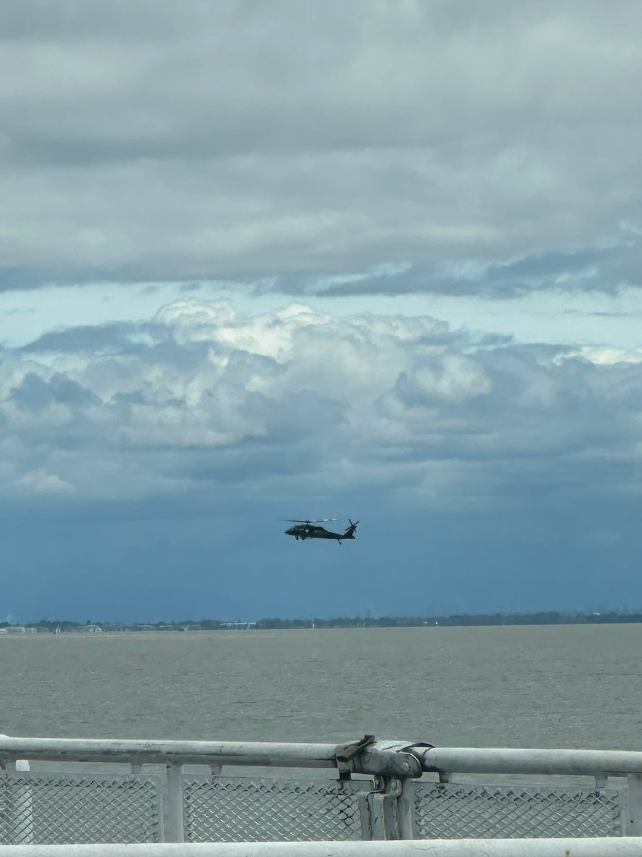 A black helicopter hovers over the ocean with the railing of a ferry in the foreground.