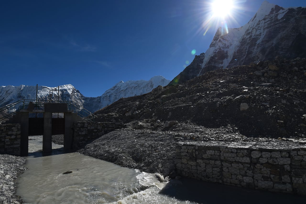 Water flows through a rock and concrete canal in front of tall snowcapped mountains on a clear, sunny day.