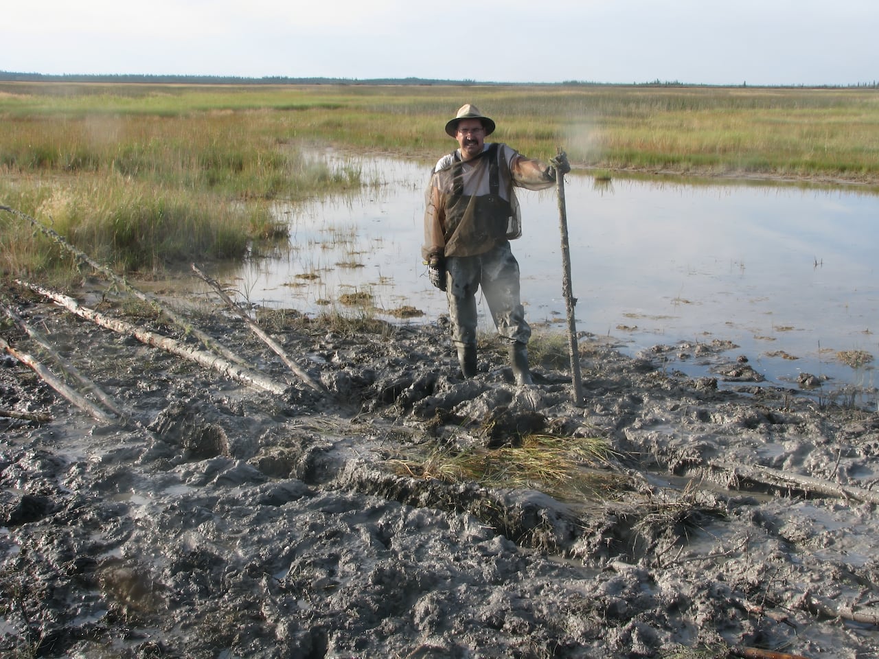 A man wearing a hat and wellington boots stands in a marsh while holding a walking stick