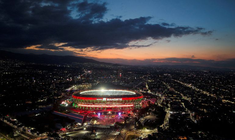 Azteca Stadium lit up as the sun sets with dark clouds overhead