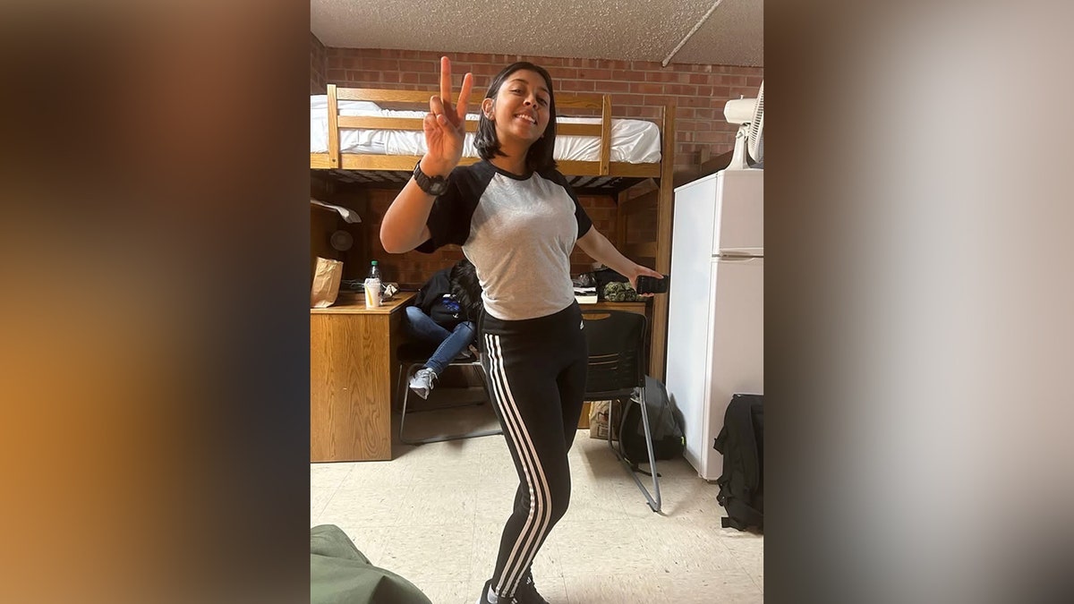 Angelina Resendiz smiles and flashes a peace sign while standing inside a Navy barracks room.
