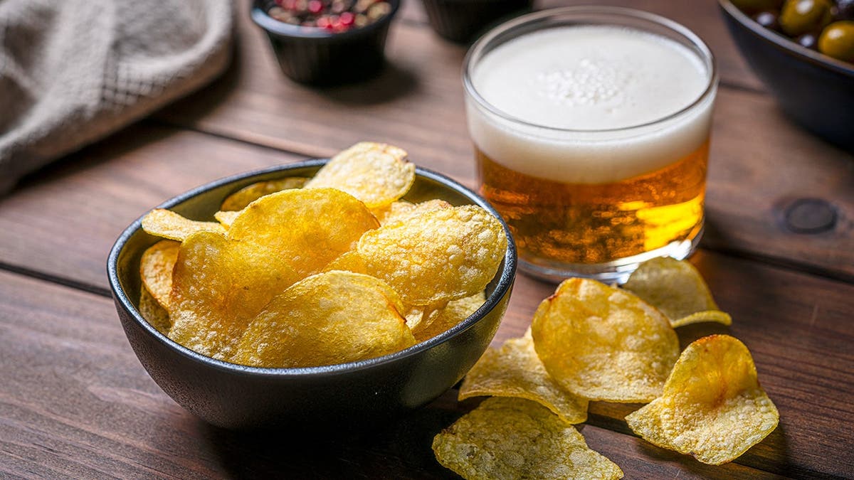 A bowl of chips sits on a table beside a glass of beer.
