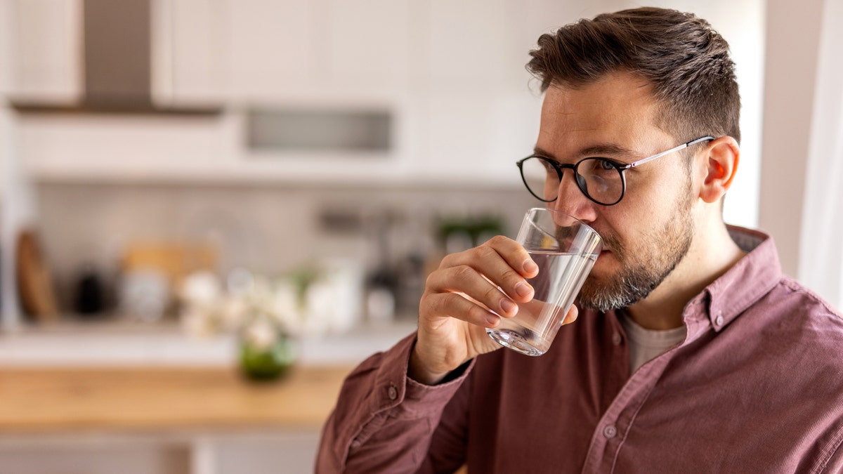 A thoughtful man drinking a glass of water in a home kitchen.