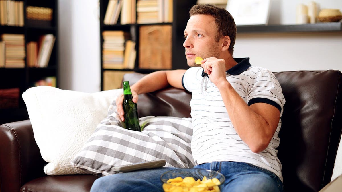 A man sitting on a couch holds a beer bottle and eats from a bowl of chips.
