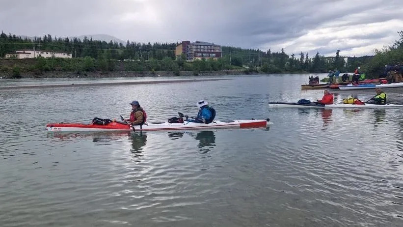 Ian Tyers and Jim Nye are in a canoe on the River Yukon. They are wearing life jackets and hats and there are several other canoes on the water behind them. There are several buildings in the distance and fir trees are growing near the far river bank.