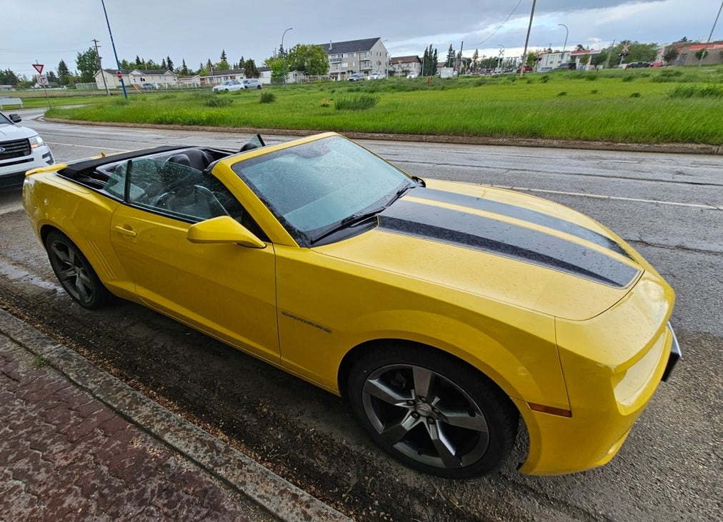 A yellow car with black stripes down the centre is parked on the side of the road.