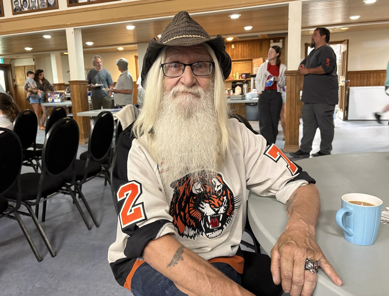 A man in a hat and long white beard sits in a room where people are gathered.