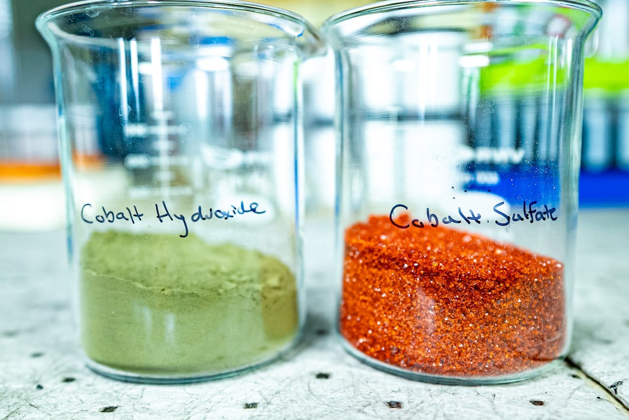 Image shows two glass beakers side by side in a lab. One beaker has green powder and the other beaker has reddish crystals.