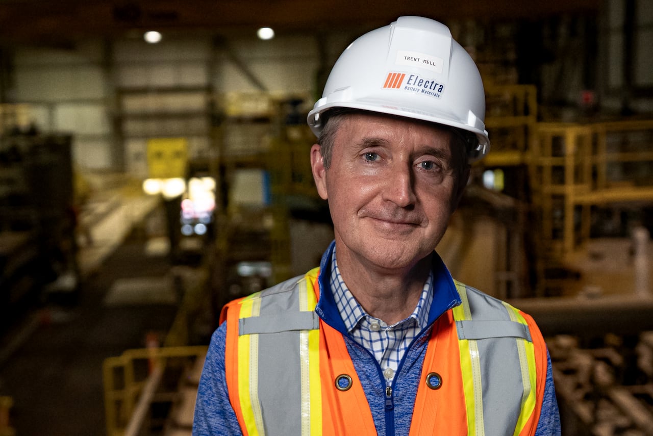 Man in white hard hat and orange safety vest smiling at the camera and standing in  a factory-type setting.