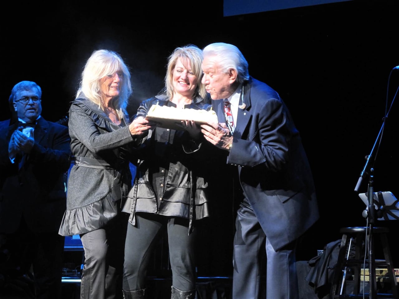 A man blows out a candle on a birthday cake. Two women also hold the cake.